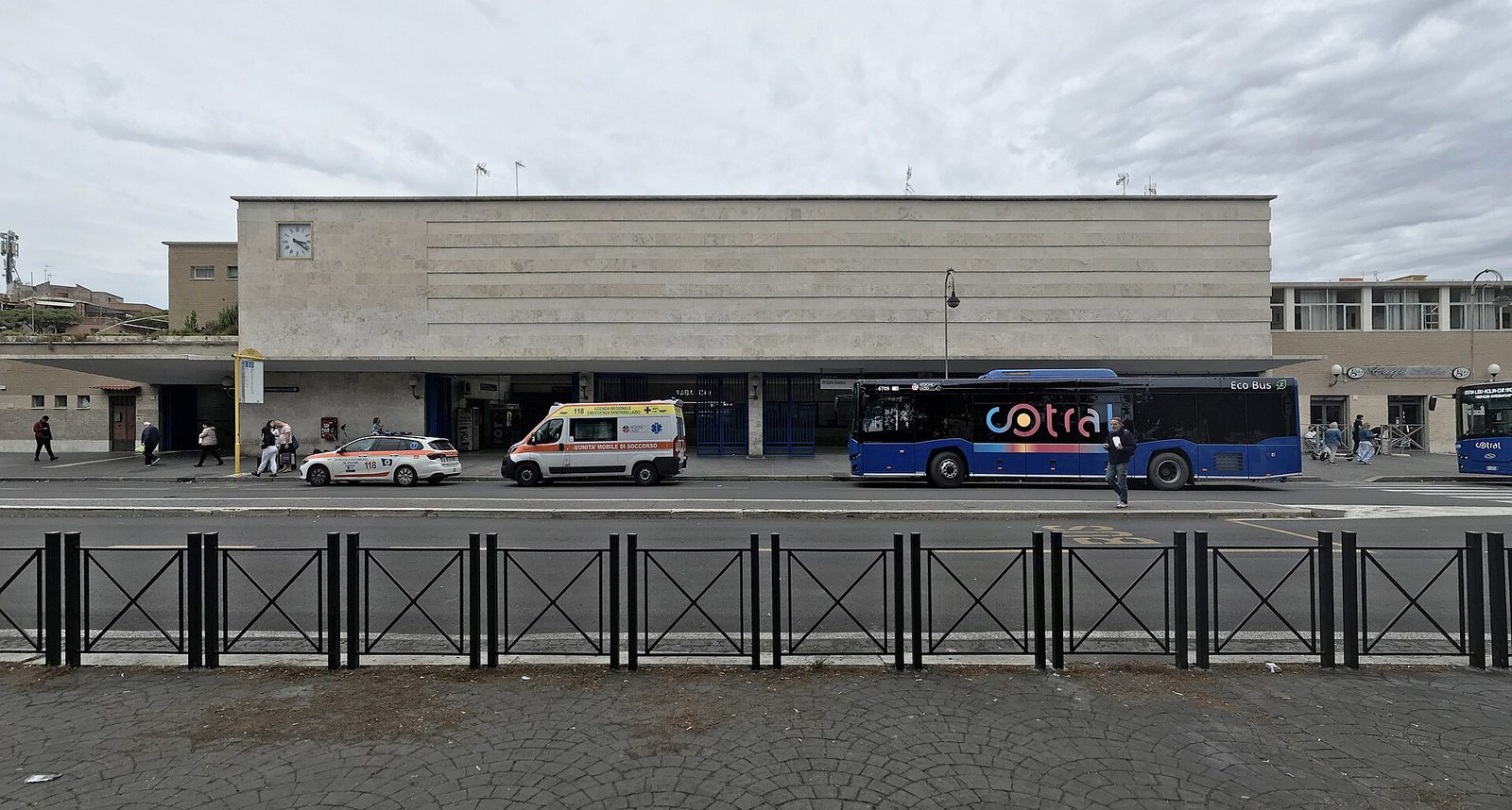 La facciata della stazione Lido Centro a Ostia: un lungo edificio rivestito in travertino con orologio, la pensilina sull'ingresso e un autobus blu Cotral fermo davanti, sotto un cielo nuvoloso
