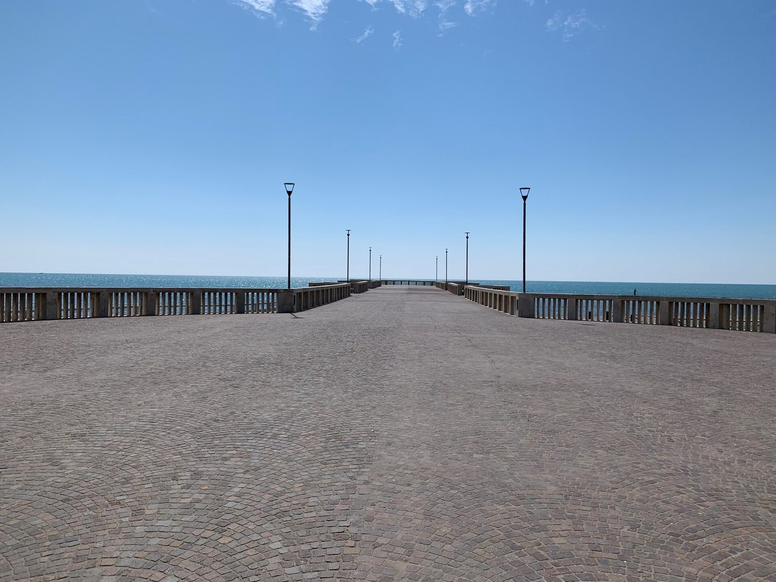Il Pontile di Lido di Ostia visto dal piazzale d'ingresso: una lunga passerella lastricata a cubetti, con balaustre chiare e lampioni, che si allunga verso il mare blu sotto un cielo sereno