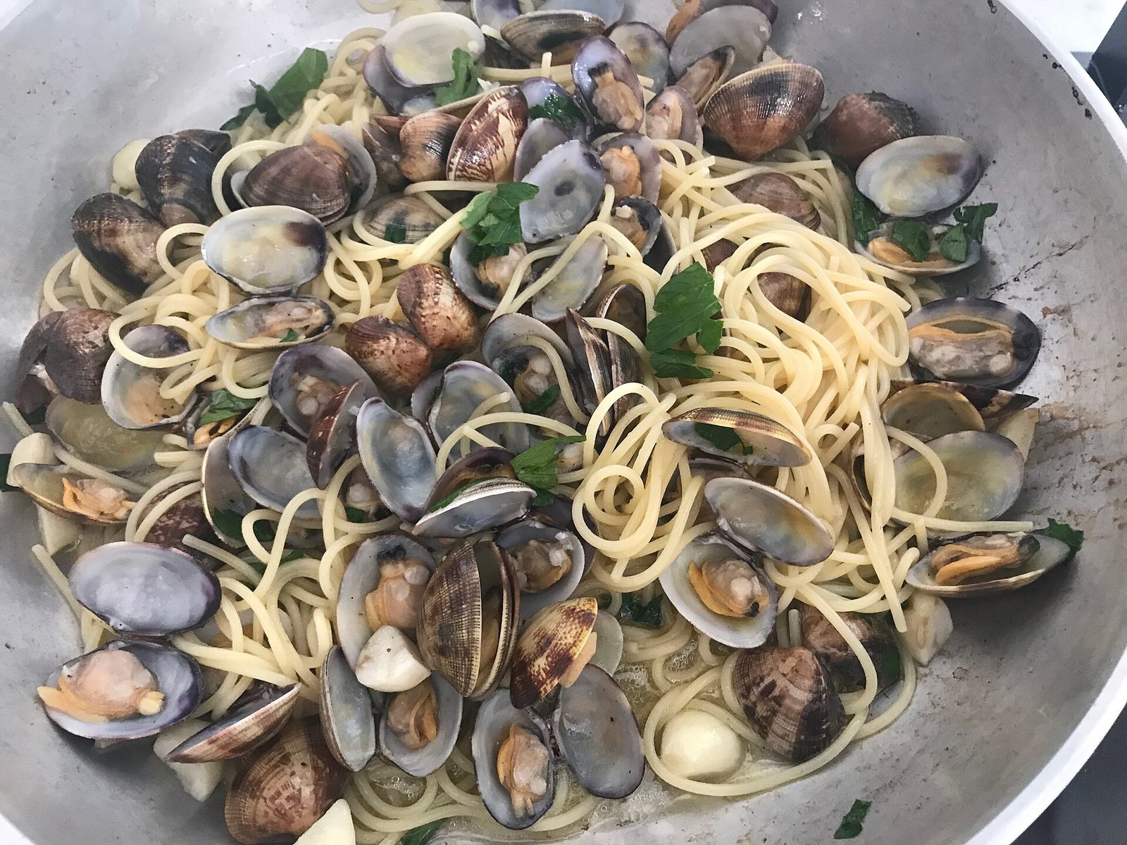 Una padella di alluminio piena di spaghetti saltati con vongole aperte, spicchi d'aglio e prezzemolo fresco tritato