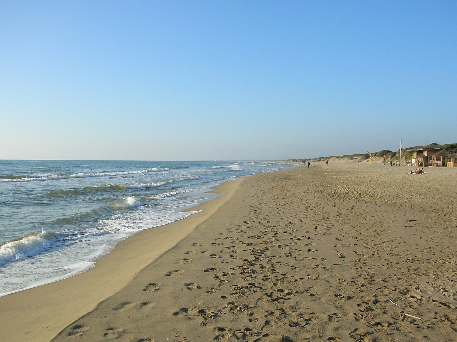 La lunga spiaggia di Capocotta: sabbia dorata segnata da impronte, onde che si infrangono sulla riva a sinistra e, sulla destra, le dune coperte di vegetazione con alcuni capanni in legno e pochi passanti in lontananza